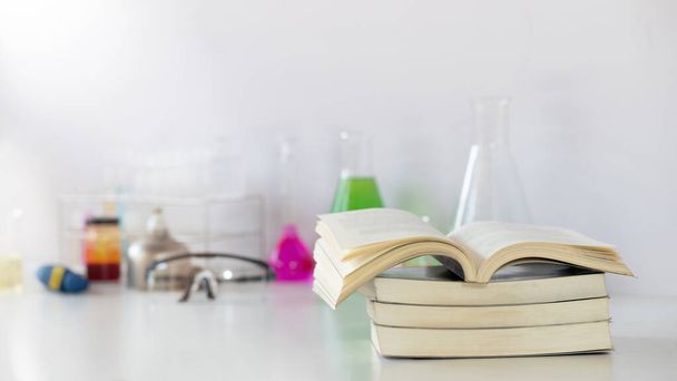 Attrezzature scientifiche, vetreria chimica e pila di libri che mettono insieme su una scrivania bianca su una parete bianca di laboratorio come sfondo. - Foto, immagini
