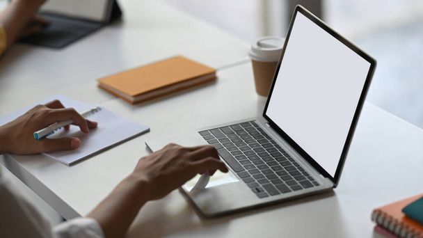Przycięty obraz rąk sekretarki piszącej na laptopie komputerowym z białym, czystym ekranem, który umieszcza na uporządkowanym biurku roboczym otoczonym notatkami, filiżanką kawy i stosem książek. - Zdjęcie, obraz