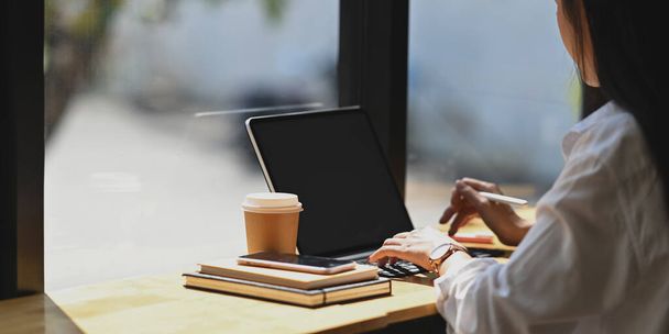 Image latérale de la belle femme d'affaires en chemise blanche tapant sur la tablette d'ordinateur avec clavier tout en étant assis à la table de travail en bois avec des fenêtres de café / restaurant comme arrière-plan. - Photo, image