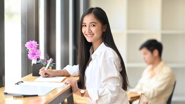 Photo of young beautiful woman working as accountant writing on document file while sitting at the wooden counter bar with colleague in office as background. - Photo, Image