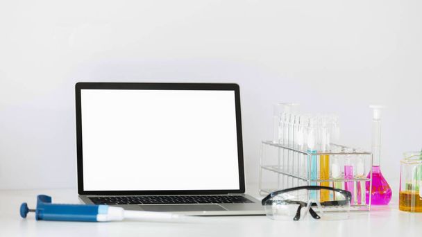 Φωτογραφία του επιστημονικού εξοπλισμού πειραμάτων που βάζει στο άσπρο γραφείο εργασίας με το άσπρο κενό lap-top υπολογιστών οθόνης. Επίπεδος lay laptop υπολογιστή, γυαλί χημείας, γυαλιά ασφαλείας. - Φωτογραφία, εικόνα