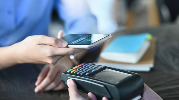 Zugeschnittenes Bild von Smart-Man-Händen, die ein Smartphone in der Hand halten und mit einer NFC-Technologie am Kreditkartenlesegerät bezahlen, das den Café-Bezahlschalter aufsetzt. Technologie- und Zahlungskonzept. - Foto, Bild