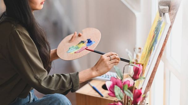 Обрізане зображення молодої дівчини-художниці, яка тримає пензлик і малює кольори олії на полотні, сидячи в сучасній художній студії. Концепція творчої жінки як художника
. - Фото, зображення