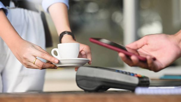Przycięty obraz rąk inteligentnego człowieka trzymających smartfona i dokonujących płatności za pomocą technologii NFC w czytniku kart kredytowych, który stawia na licznik płatności kawiarni. Technologia i koncepcja płatności. - Zdjęcie, obraz