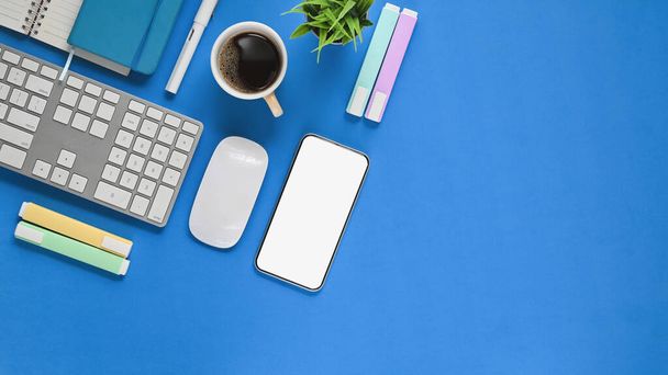 Top view image of workspace. Flat lay white blank screen smartphone, coffee cup, pen, marker pens, notebook, keyboard, wireless mouse and potted plant on blue working table. - Photo, Image