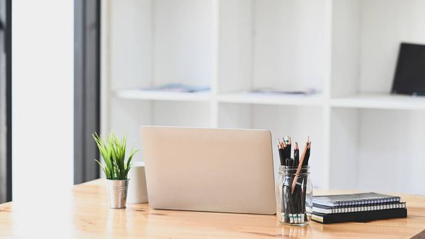 Computer-Laptop, Bleistifthalter, Topfpflanze, Notizbuch und Kaffeetasse auf modernem Holztisch mit Bürobuchregal als Hintergrund. - Foto, Bild