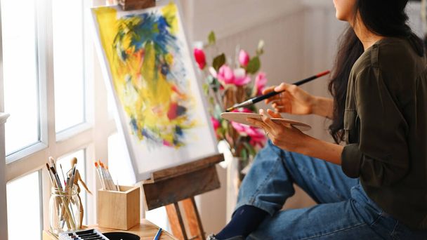 Zugeschnittenes Bild einer jungen schönen Künstlerin, die einen Pinsel hält und auf einer Leinwand zeichnet, während sie neben den Kunstaccessoires sitzt, die auf einem kleinen Holztisch mit Atelierfenstern als Hintergrund stehen.. - Foto, Bild