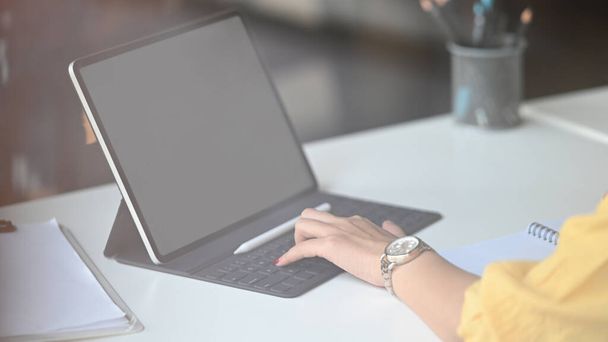 Przycięty obraz kreatywnej kobiety trzymającej pióro rysika podczas rysowania / wskazywania na biały pusty ekran komputera tablet z klawiaturą, że umieszczanie na biurku pracy na wygodnym pomieszczeniu jako tło. - Zdjęcie, obraz