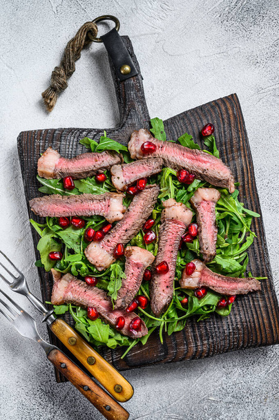 Gesneden gegrilde biefstuk met arugula bladeren salade op rustieke snijplank. Witte achtergrond. Bovenaanzicht - Foto, afbeelding
