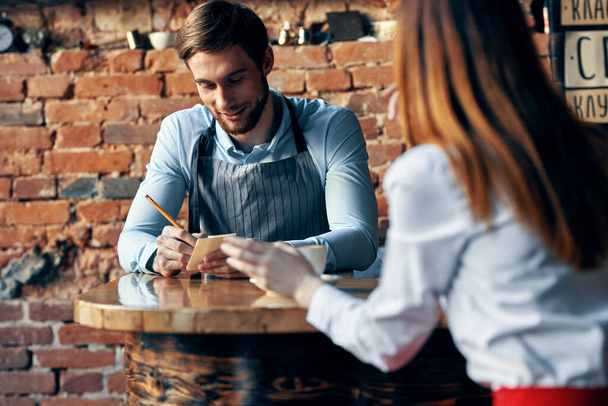 мужчина официант в серых фартуках принимает заказ и чашку кофе женщина клиент за столом в кафе - Фото, изображение