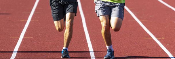 Vista frontal de las piernas de dos corredores corriendo en carriles uno al lado del otro en una pista roja bajo el sol brillante. - Foto, Imagen
