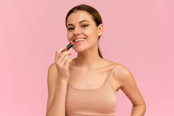 Μια νεαρή γυναίκα βάζει γυμνό κραγιόν στα χείλη της σε ροζ φόντο. Έννοια καλλυντικών - Φωτογραφία, εικόνα