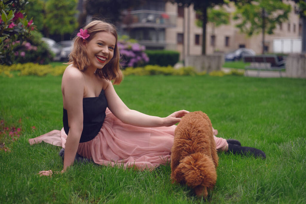 A girl in a long pink dress is sitting in the yard with her dog, a brown poodle. The girl walks with the dog, the hostess and the animal. A girl is playing with her dog. - 写真・画像