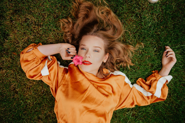 A girl with healthy beautiful hair lies on the grass and looks at the camera. Girl with perfect skin and natural care. Flower in the mouth holds a pink flower near the face. Natural beauty, youth, teenager. Red lips - Фото, изображение