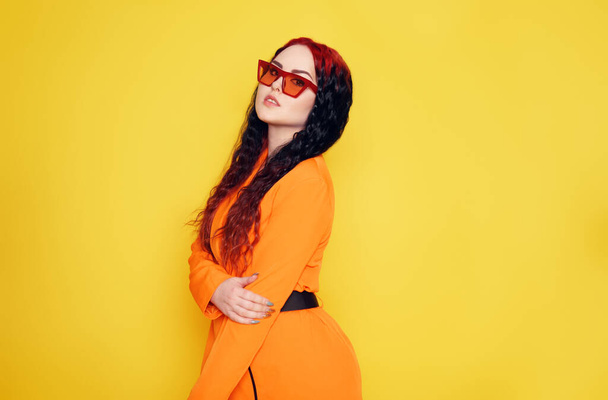 Beautiful girl on a yellow background. A brunette in an orange dress and long hair is posing, smiling, having fun. Woman in sunglasses. Joy and emotion. Red hair roots place for text - Photo, Image