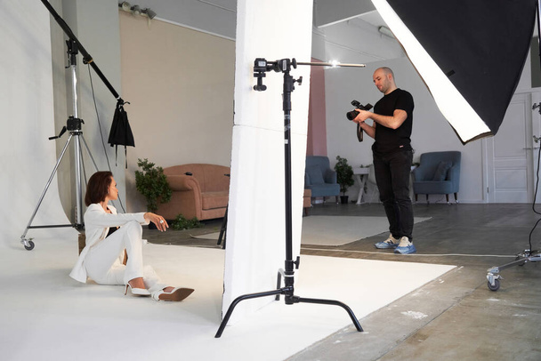 Fashion photography in a photo studio. Professional male photographer taking pictures of beautiful woman model on camera, backstage - Photo, image