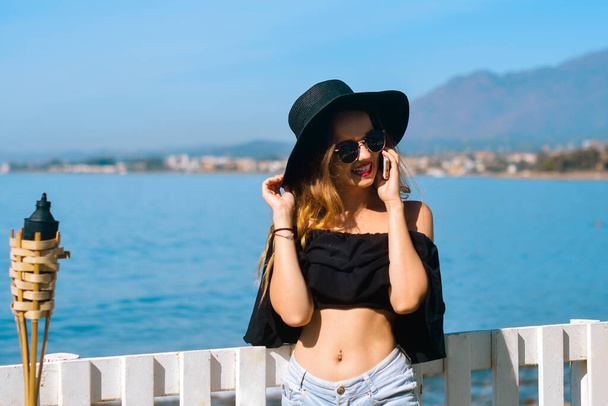 A beautiful girl speaks on the phone against the backdrop of the beach and ocean, sea. Woman in a big black hat and glasses on vacation. Freelancer calls and smiles, roaming and chatting. Travel - Foto, imagen