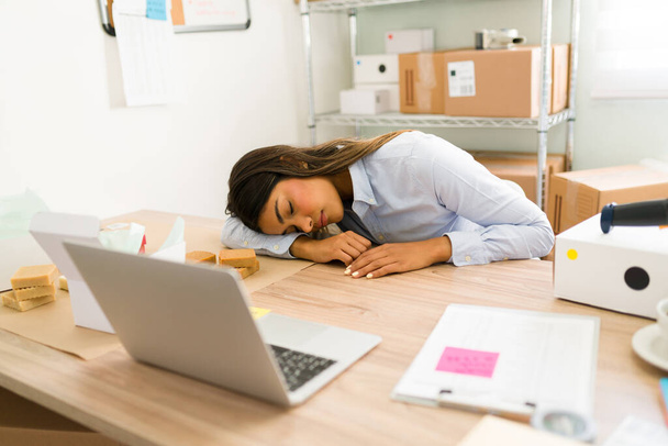 Väsynyt ahkera nuori nainen ja yrityksen omistaja nukkuu toimistossaan työpöydän ääressä kiireisen päivän jälkeen kauneustuotteiden verkkokaupassa - Valokuva, kuva