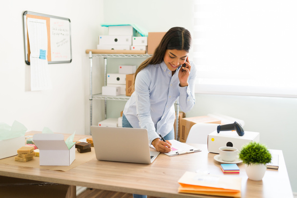 Wesoła kobieta przedsiębiorca rozmawia przez telefon z klientem lub jej dostawcą i zapisuje informacje podczas pracy w swojej firmie  - Zdjęcie, obraz