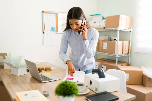 Εργατική νεαρή γυναίκα μιλάει στο τηλέφωνο, ενώ δίνει εξυπηρέτηση πελατών για τις online παραγγελίες και πακέτα της από το κατάστημα της επιχείρησής της - Φωτογραφία, εικόνα