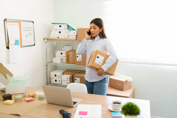 Providing customer service. Good-looking young woman talking on the phone with a customer and answering questions while arranging packages ready to ship - Photo, image