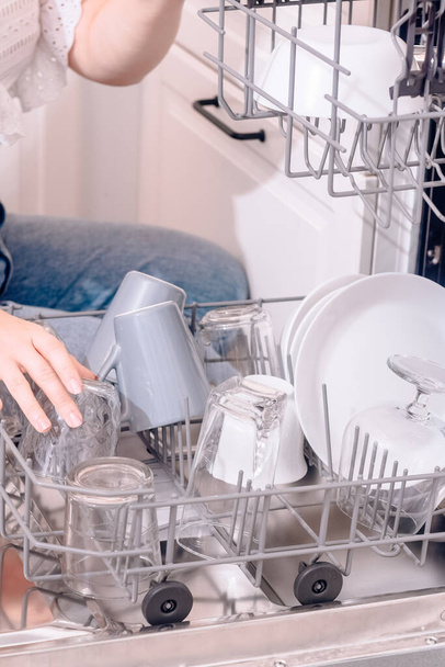 Lava-louças embutida, lava-louças. Uma mulher carrega pratos lavados, copos, copos. A mão gentil de uma mulher coloca algo na máquina de lavar louça ou puxa, descarrega. - Foto, Imagem