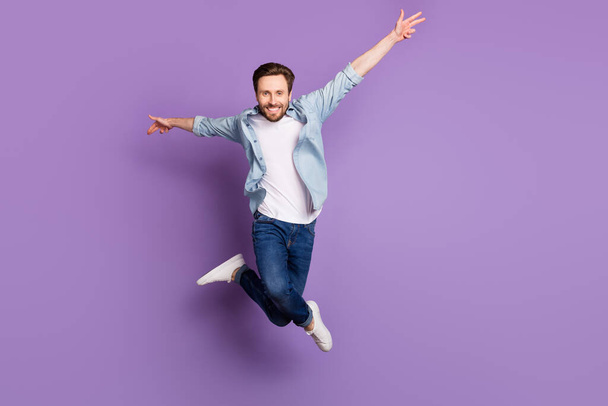 Photo of excited funny young man wear jeans shirt jumping high rising arms hands sides isolated violet color background - Foto, afbeelding
