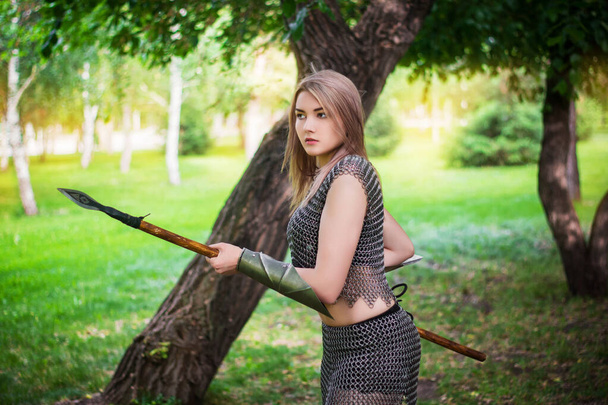 Portrait of a young medieval woman warrior dressed in chain mail armor with a spear in her hands on the background of the forest. - Foto, imagen