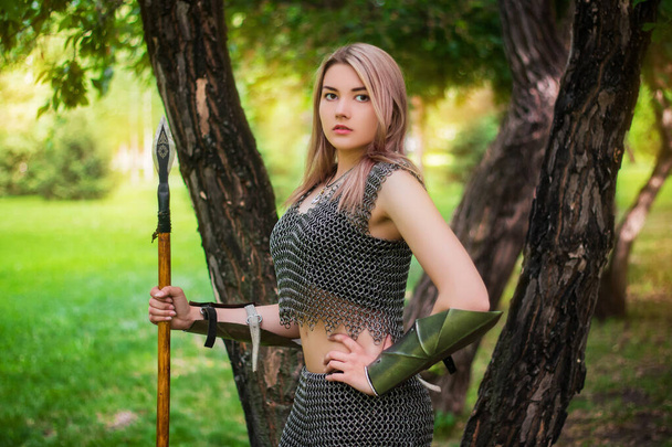 Portrait of a young medieval woman warrior dressed in chain mail armor with a spear in her hands on the background of the forest. - Fotó, kép