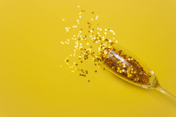 Las estrellas de Confetti se derraman de una copa de champán sobre un fondo amarillo. La vista desde arriba. El concepto de la celebración. - Foto, imagen