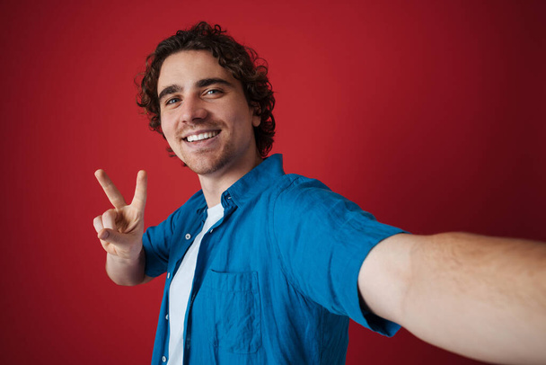 Joyful handsome man showing peace sign and taking selfie photo isolated over red background - Fotografie, Obrázek