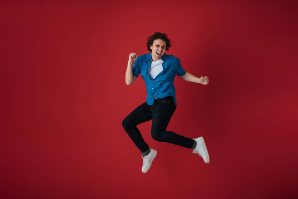 Excited handsome young man making fun while jumping isolated over red background - Фото, зображення