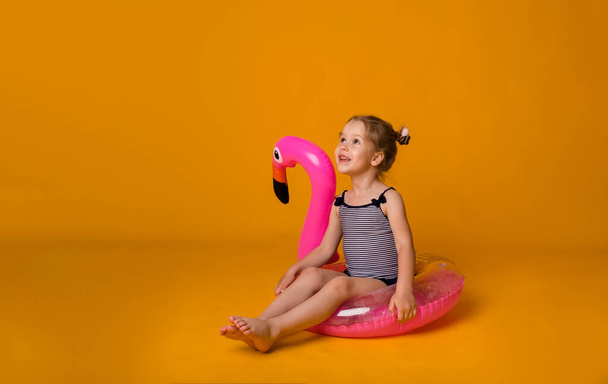 little blonde girl in a striped swimsuit sits in a pink inflatable circle with a flamingo and looks away on a yellow background - Fotografie, Obrázek
