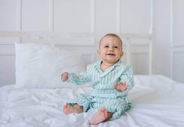 vrolijk jongetje in gestreepte pyjama zit op het witte beddengoed op het bed en kijkt in de camera. Gelukkige kindertijd - Foto, afbeelding