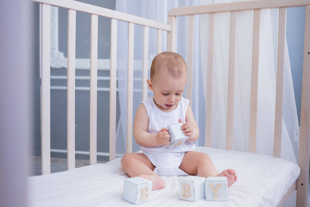 Beyaz elbiseli akıllı küçük çocuk, elinde tahta küplerle bir çocuğun yatağında oturuyor. Erken çocuk gelişimi - Fotoğraf, Görsel