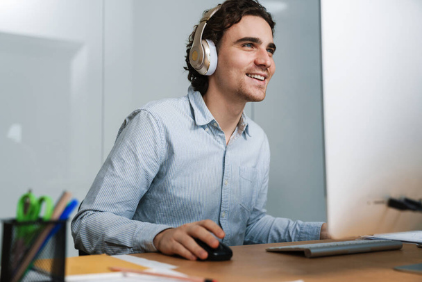 Caucasian young happy businessman in headphones smiling while working on computer in office - 写真・画像