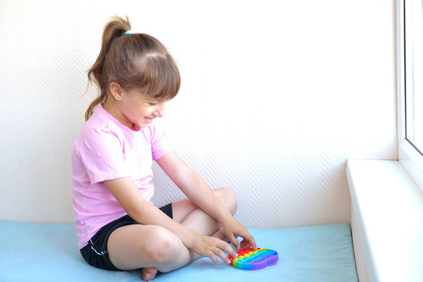 a girl in a pink t-shirt plays with a trendy toy pop it. colorful antistress sensory toy fidget push pop it in kid's hands, can be used for training with autistic people - Photo, Image