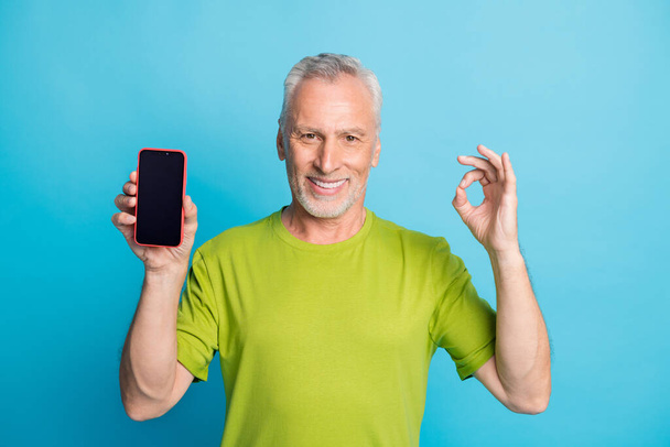 Portrait of attractive cheerful grey-haired man showing ok-sign holding in hand device isolated over bright blue color background - Photo, image