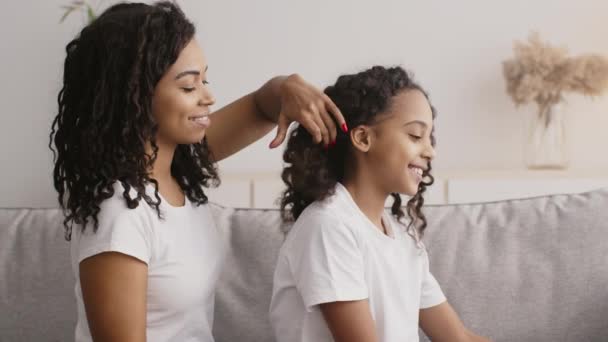 Mladá veselá africká americká dáma kartáčování vlasy své roztomilé kudrnaté dcerušky, sedí spolu na pohovce doma - Záběry, video