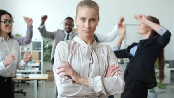 Serious businesswoman with blond hair standing in office while coworkers dancing in background - Materiaali, video
