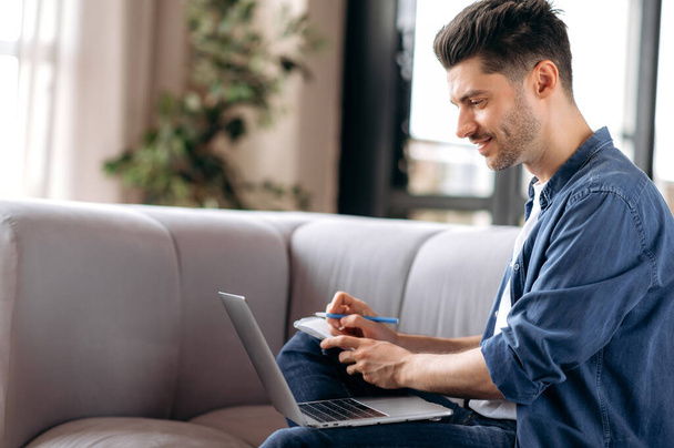 Concentrated young adult Caucasian man, freelancer or student, in casual clothes, sitting on sofa in living room, using laptop for video calling with colleagues, looking at screen, taking notes - Photo, Image