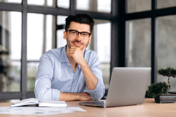 Portrait handsome young caucasian freelancer or manager, wearing strict shirt and glasses, sitting at table with laptop, supporting his head with his hand, looking at camera and smiling friendly - Фото, изображение