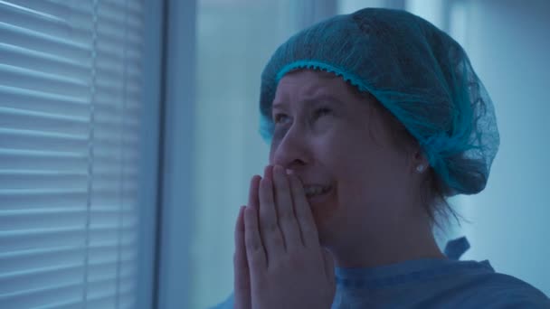 Gebed voor de gezondheid. Verzoek aan God voor de operatie. Vrouw stress en huilen na een ongeval met gelijke kaak, tanden en gezicht bidden met gevouwen handen in de buurt van haar gezicht, terwijl staan door het raam in het ziekenhuis - Video