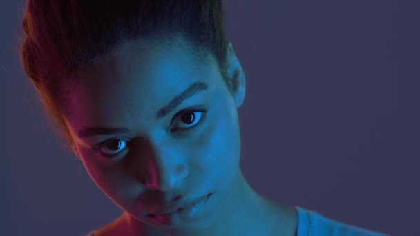 Jovem afro-americana sombria olhando sullenly para câmera, posando em luzes de néon fundo, close up retrato - Filmagem, Vídeo