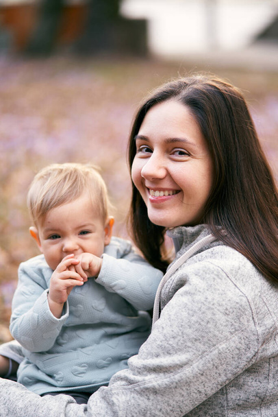 笑顔のお母さんは公園の葉の上に座っている間、彼の口の中で手で幼児を抱きます。接近中だ。肖像画 - 写真・画像