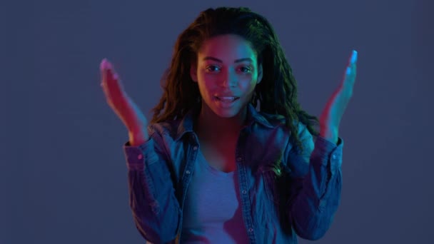 Giovane donna afroamericana che apre il viso e si sente stupita, stendendo le mani nello smarrimento nella luce al neon - Filmati, video