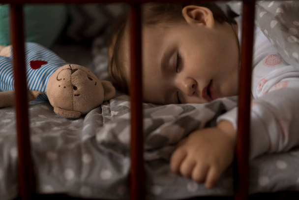 Jolie petite fille joufflue et joyeuse qui dort doucement dans un berceau gris pendant la pause déjeuner en pyjama blanc et rose avec ours en peluche à la maison. Enfance, loisirs, confort, médecine, concept de santé - Photo, image