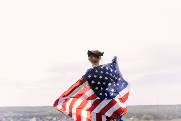 Young beautiful woman holding USA flag. The back of a young woman with the US national flag on her shoulders against the backdrop of the mountains celebrates the Independence Day of the United States - Foto, Imagen