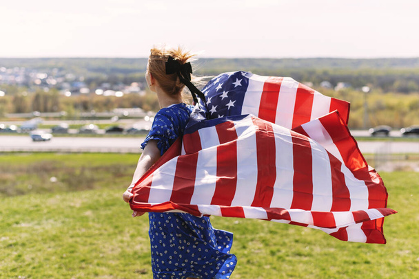 A young woman with the national flag of the USA on her shoulders against the backdrop of the city celebrates the Independence Day of the United States - Fotografie, Obrázek
