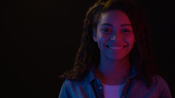 Junge fröhliche Afroamerikanerin lächelt in Neonlichter in die Kamera, schwarzer Hintergrund mit leerem Raum - Filmmaterial, Video